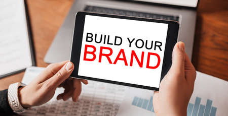 Man hands holding tablet with text Build Your Brand at workplace. Businessman working at desk with documentsの写真素材