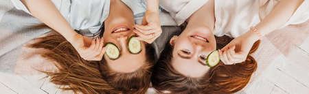 Two girls make homemade face and hair beauty masks. Cucumbers for the freshness of the skin around the eyes. Women take care of youthful skin. Girlfriends laugh at home lying on the floor on pillows.の写真素材