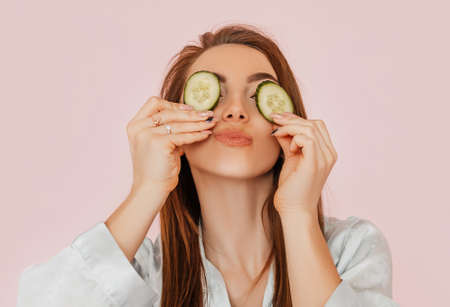 Girl make homemade face beauty masks. Cucumbers for the freshness of the skin around the eyes. Woman take care of youthful skin. Model laughing and having fun in spa on pink backgroundの写真素材