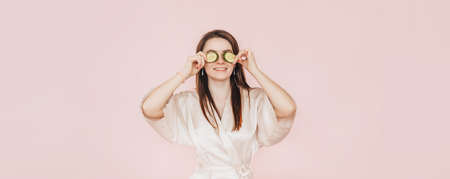Girl make homemade face beauty masks. Cucumbers for the freshness of the skin around the eyes. Woman take care of youthful skin. Model laughing and having fun in spa on pink backgroundの写真素材