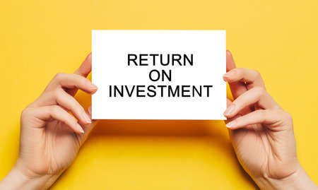 Female hands hold card paper with text Return on Investment on a yellow background. Business and finance conceptの写真素材