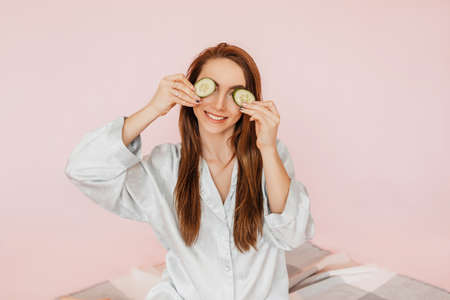 Girl make homemade face beauty masks. Cucumbers for the freshness of the skin around the eyes. Woman take care of youthful skin. Model laughing and having fun in spa on pink backgroundの写真素材