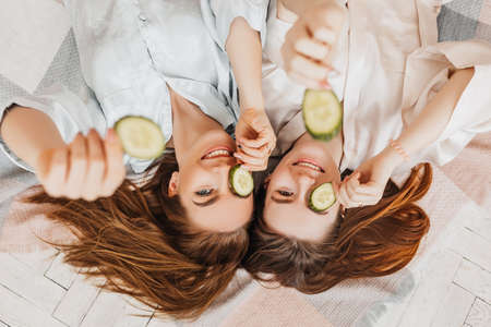 Two girls make homemade face and hair beauty masks. Cucumbers for the freshness of the skin around the eyes. Women take care of youthful skin. Girlfriends laugh at home lying on the floor on pillows.の写真素材