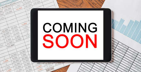 Tablet with text COMING SOON on your desktop with documents, reports and graphs. Business and finance conceptの写真素材
