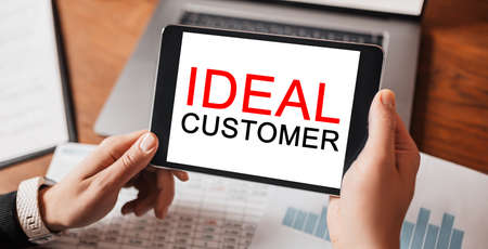 Man hands holding tablet with text IDEAL CUSTOMER at workplace. Businessman working at desk with documentsの写真素材