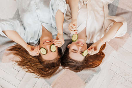 Two girls make homemade face and hair beauty masks. Cucumbers for the freshness of the skin around the eyes. Women take care of youthful skin. Girlfriends laugh at home lying on the floor on pillows.の写真素材