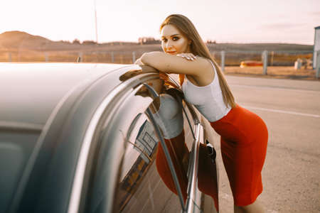 Beautiful young girl near a car in the evening in the sunset sun in an empty parking lotの写真素材