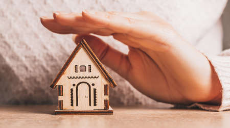 Woman holds and protects a wooden house with her hands with the sun on a light pink background. Sweet homeの写真素材