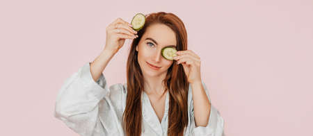Girl make homemade face beauty masks. Cucumbers for the freshness of the skin around the eyes. Woman take care of youthful skin. Model laughing and having fun in spa on pink backgroundの写真素材