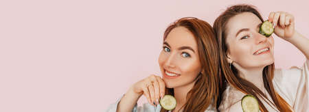 Two girls make homemade face beauty masks. Cucumbers for the freshness of the skin around the eyes. Women take care of youthful skin. Girlfriends laughing and having fun in spa on pink backgroundの写真素材