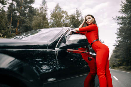 A beautiful young girl in a red overalls stands by a black car on an empty road in the forestの写真素材