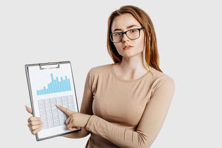 Beautiful young business woman wearing glasses isolated on gray background. Achieving career wealth business concept. Mockup copy space. Hold a clipboard with documents charts and reportsの写真素材