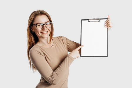 Beautiful young business woman wearing glasses holds a clipboard with mock up blank space isolated on gray background. Achieving career wealth business concept. Online learning coverの写真素材
