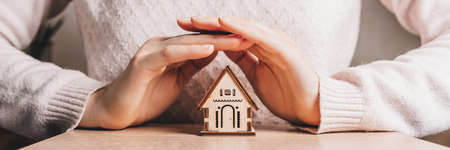 Woman holds and protects a wooden house with her hands with the sun on a light pink background. Sweet homeの写真素材