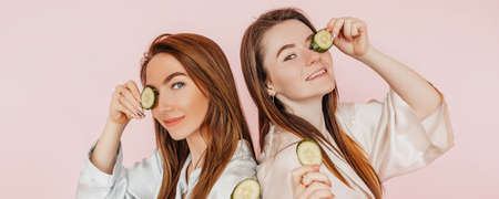 Two girls make homemade face beauty masks. Cucumbers for the freshness of the skin around the eyes. Women take care of youthful skin. Girlfriends laughing and having fun in spa on pink backgroundの写真素材