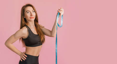 Fitness girl smiling and posing hold measure tape in black sportswear on a pink background. Slim woman with a beautiful athletic body and tanned skinの写真素材