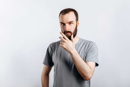 Serious puzzled handsome male model with beard holding hand on chin as if thinking about something, squinting at camera with suspicious look and standing over gray background. Man decides what to buyの写真素材