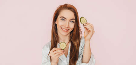 Girl make homemade face beauty masks. Cucumbers for the freshness of the skin around the eyes. Woman take care of youthful skin. Model laughing and having fun in spa on pink backgroundの写真素材