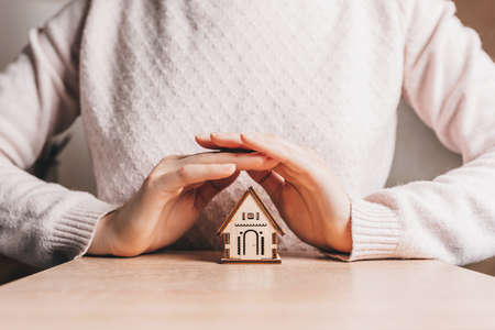 Woman holds and protects a wooden house with her hands with the sun on a light pink background. Sweet homeの写真素材