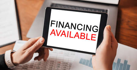 Man hands holding tablet with text Financing Available at workplace. Businessman working at desk with documentsの写真素材