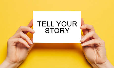Female hands hold card paper with text Tell Your Story on a yellow background. Business and finance conceptの写真素材