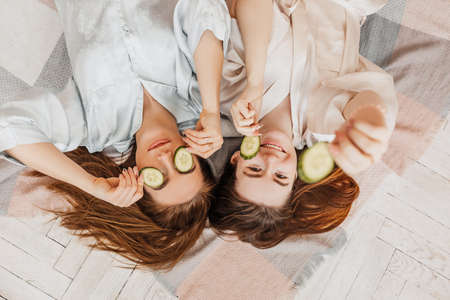 Two girls make homemade face and hair beauty masks. Cucumbers for the freshness of the skin around the eyes. Women take care of youthful skin. Girlfriends laugh at home lying on the floor on pillows.の写真素材