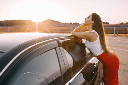 Beautiful young girl near a car in the evening in the sunset sun in an empty parking lotの写真素材