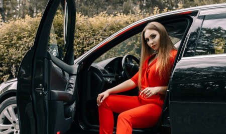 A beautiful young girl in a red overalls sits behind the wheel of a black car on an empty road in the forestの写真素材