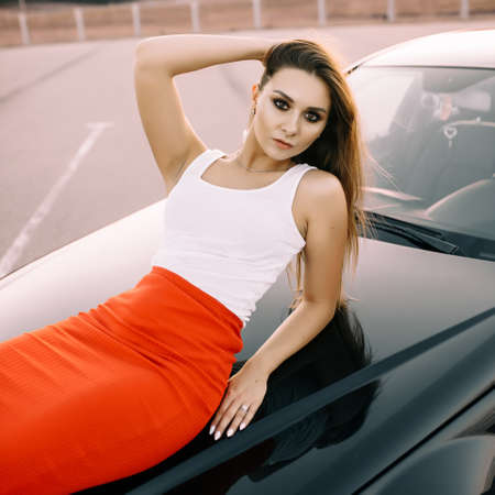 Beautiful young girl near a car in the evening in the sunset sun in an empty parking lot