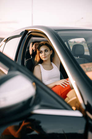 A beautiful young girl sits in a car in the evening in the sunset summer sun in an empty parking lotの写真素材
