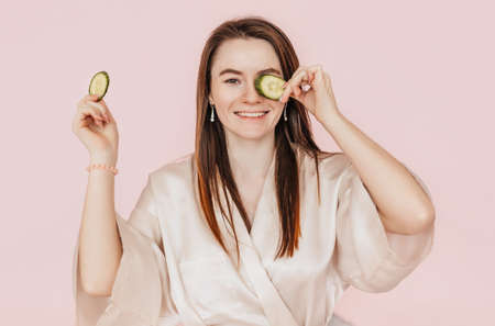 Girl make homemade face beauty masks. Cucumbers for the freshness of the skin around the eyes. Woman take care of youthful skin. Model laughing and having fun in spa on pink backgroundの写真素材