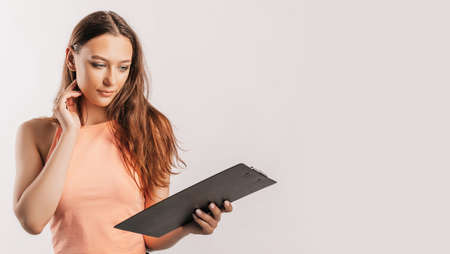 Girl student writes information. Beautiful young business woman isolated on gray background. Achieving career wealth business concept. Mockup copy space. Hold a clipboard with documentsの写真素材