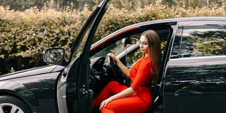 A beautiful young girl in a red overalls sits behind the wheel of a black car on an empty road in the forestの写真素材