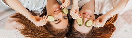Two girls make homemade face and hair beauty masks. Cucumbers for the freshness of the skin around the eyes. Women take care of youthful skin. Girlfriends laugh at home lying on the floor on pillows.の写真素材