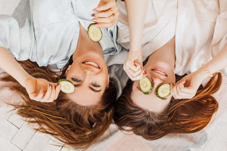 Two girls make homemade face and hair beauty masks. Cucumbers for the freshness of the skin around the eyes. Women take care of youthful skin. Girlfriends laugh at home lying on the floor on pillows.の写真素材