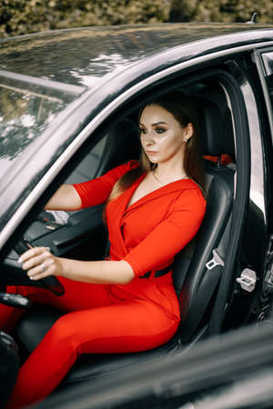 A beautiful young girl in a red overalls sits behind the wheel of a black car on an empty road in the forestの写真素材