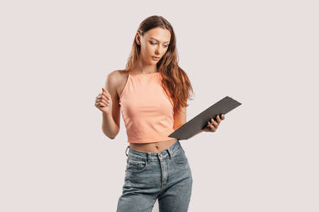 Girl student writes information. Beautiful young business woman isolated on gray background. Achieving career wealth business concept. Mockup copy space. Hold a clipboard with documentsの写真素材
