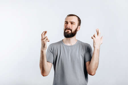 Handsome young man frown and cross fingers in hope on white background with space for advertising mock upの写真素材