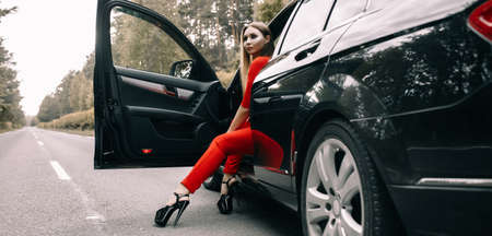 A beautiful young girl in a red overalls sits behind the wheel of a black car on an empty road in the forestの写真素材