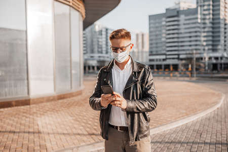 Handsome man in a protective mask on his face with glasses with a smartphone on the street of a big city. Businessman talking on the phone on urban backgroundの写真素材