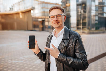 Handsome man in glasses with a smartphone on the street of a big city. Businessman points finger at phone screen on urban backgroundの写真素材