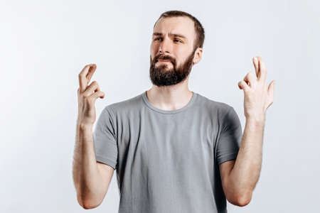Handsome young man frown and cross fingers in hope on white background with space for advertising mock upの写真素材