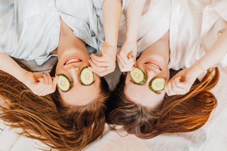 Two girls make homemade face and hair beauty masks. Cucumbers for the freshness of the skin around the eyes. Women take care of youthful skin. Girlfriends laugh at home lying on the floor on pillows.の写真素材