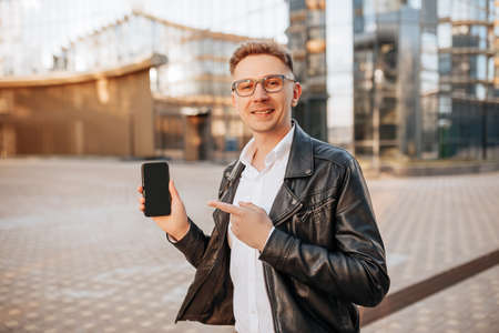 Handsome man in glasses with a smartphone on the street of a big city. Businessman points finger at phone screen on urban backgroundの写真素材
