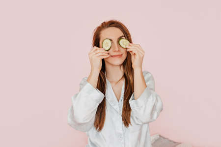 Girl make homemade face beauty masks. Cucumbers for the freshness of the skin around the eyes. Woman take care of youthful skin. Model laughing and having fun in spa on pink backgroundの写真素材