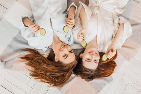 Two girls make homemade face and hair beauty masks. Cucumbers for the freshness of the skin around the eyes. Women take care of youthful skin. Girlfriends laugh at home lying on the floor on pillows.の写真素材
