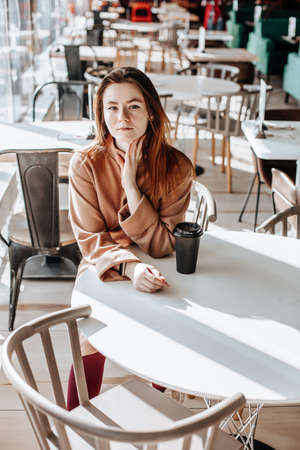 Stylish girl sits in a cafe and drinks coffee. Coffee to go in a cardboard cup. Woman with ginger hair in a beige warm suit in a cozy atmosphere. Modern interior. Calm and pleasant pastimeの写真素材