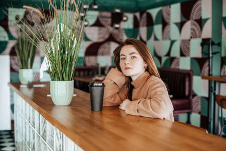Stylish girl sits in a cafe and drinks coffee. Coffee to go in a cardboard cup. Woman with ginger hair in a beige warm suit in a cozy atmosphere. Modern interior. Calm and pleasant pastimeの写真素材