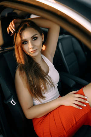 A beautiful young girl sits in a car in the evening in the sunset summer sun in an empty parking lotの写真素材