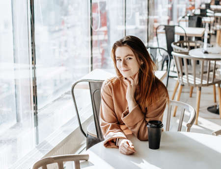 Stylish girl sits in a cafe and drinks coffee. Coffee to go in a cardboard cup. Woman with ginger hair in a beige warm suit in a cozy atmosphere. Modern interior. Calm and pleasant pastimeの写真素材